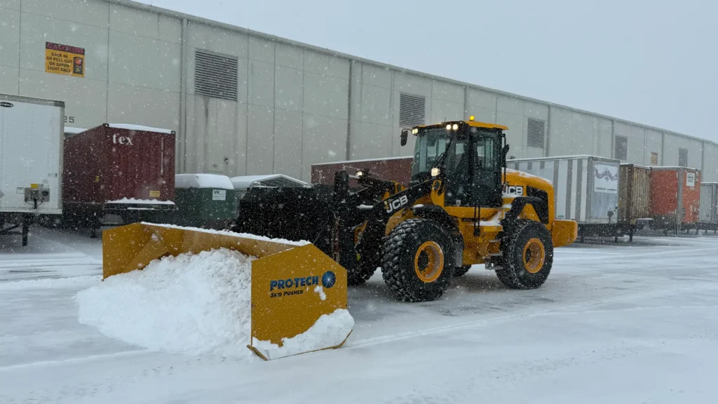 A JCB wheel loader with a Protech snow pusher clearing a commercial truck yard during a heavy snowstorm.
