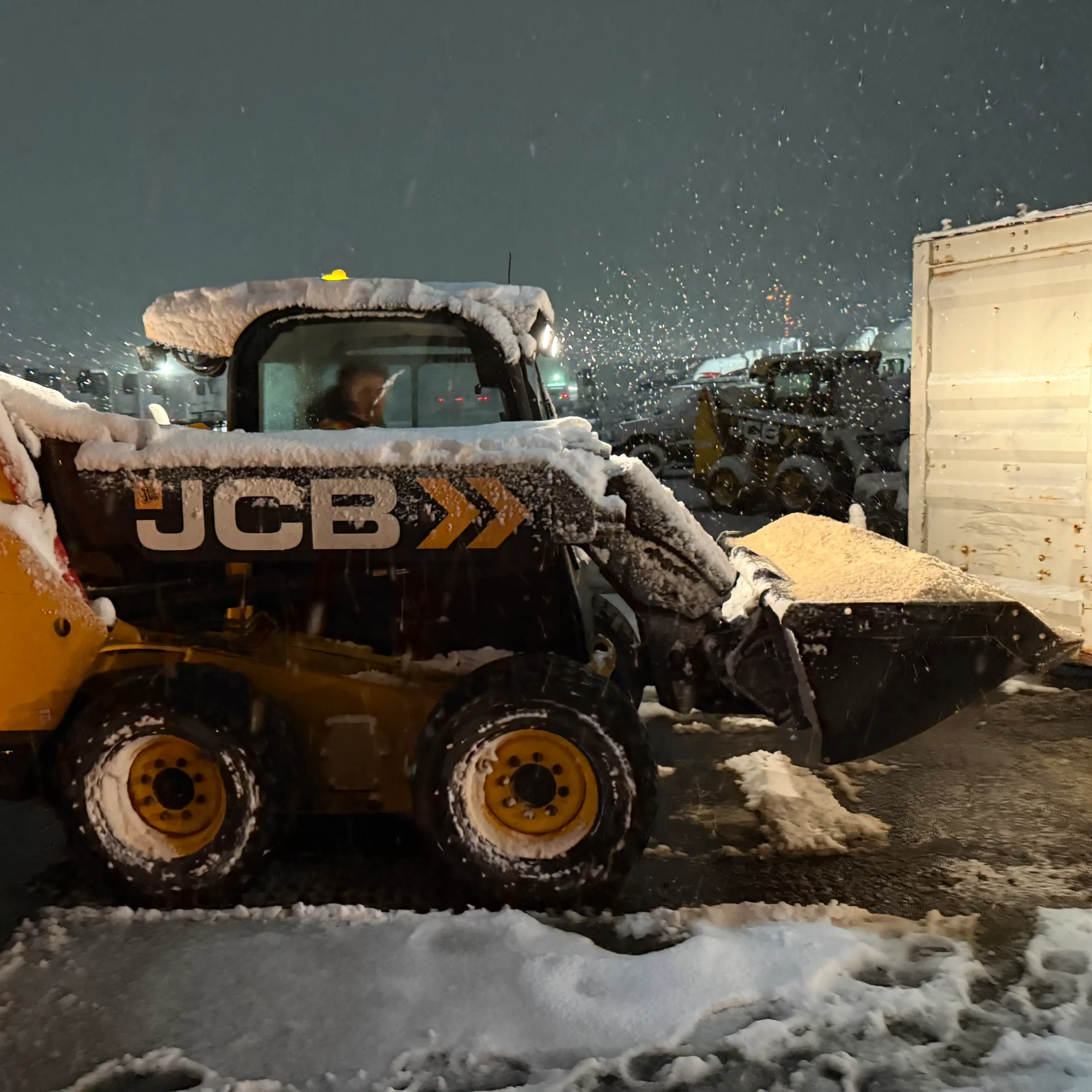 A JCB skid steer loader scooping bulk salt at night during a snowstorm in a commercial parking lot.